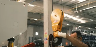 Pracownik w hali produkcyjnej sprawdza taśmę nośną przy haku suwnicy.